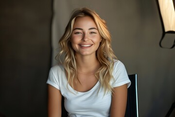 Fototapeta premium `blonde hair woman in white tshirt sitting on black chair smiling at camera, light from one side, light set up for photo shoot, beauty studio, cinematic, hyper realistic photography`