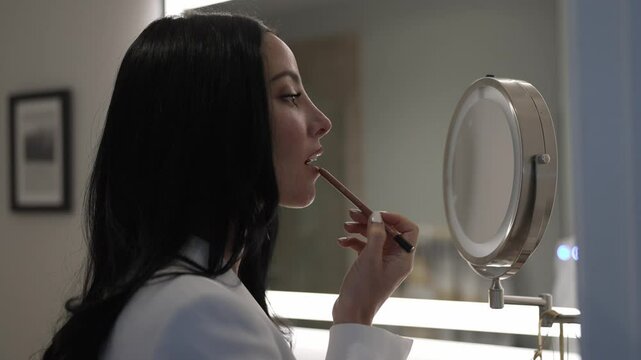 Portrait of beautiful woman applying lipliner on face, putting makeup, looking in mirror at home, getting ready in the bathroom, profile side view