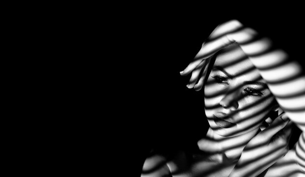 A black and white portrait of a woman in shadow. The light source casts striped shadows on her face and arm, creating a dramatic and artistic effect, copy space