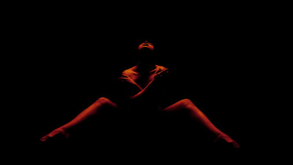 A woman is posed in a seated position, illuminated by red light. She has her arms crossed over her...