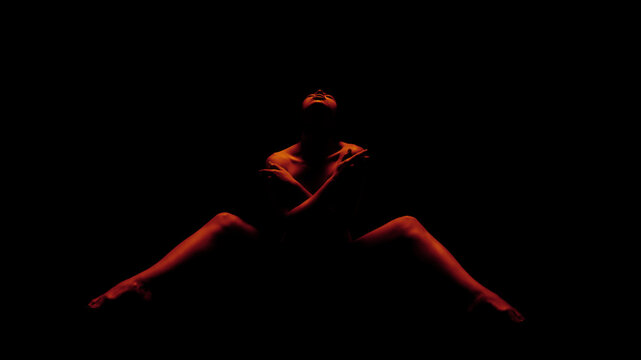 A woman is posed in a seated position, illuminated by red light. She has her arms crossed over her chest and is looking up towards the light source. The background is completely black