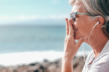 Smiling senior woman sitting in outdoors at the beach listening music by earphones, elderly female enjoying  vacation, free time and retirement
