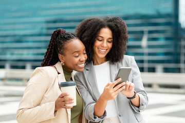 Two Black Businesswomen Smiling and Looking at Smartphone Outdoors in a Modern City