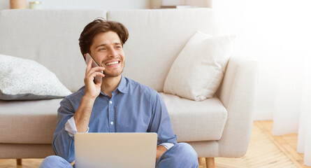 A young man sits on the floor in a living room, smiling and talking on a cell phone while working on his laptop.