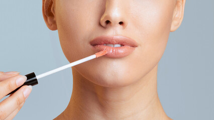 A woman with smooth skin is applying lip gloss to her lips with a clear applicator wand. The background is a light blue, cropped