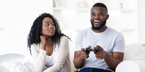 A couple sits on a couch, the man is engrossed in playing a video game, while the woman watches...