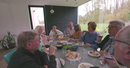 Group of elderly friends laughing and dining together at a modern, cozy table, enjoying good food and wine.