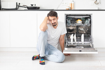 Young handsome man putting dirty plates in dishwasher machine in the kitchen. Household and...