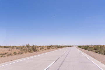 A long, empty road with a clear blue sky above. The road is mostly empty, with only a few trees in the distance. Scene is calm and peaceful, with the vast open space