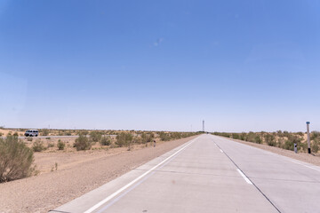 A long, empty road with a blue sky above. The road is lined with trees and there is a car driving down the road