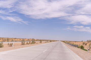 A long, empty road with a clear blue sky above. The road is lined with trees and rocks, giving it a desolate and lonely appearance