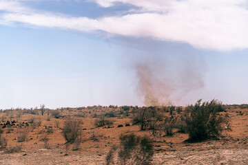 A desert landscape with a cloud of smoke in the distance. The sky is clear and blue, and the sun is shining brightly. Scene is peaceful and serene, despite the smoke in the air