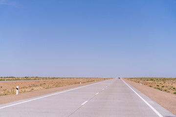 A long, empty road with a car driving down it. The sky is clear and blue, and there are no other cars or people visible