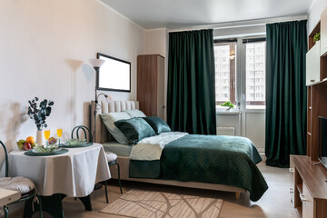 The interior of stylish bedroom with big bed, a green bedspread, green pillows and curtains, a night lamp. Near the bed there is a round table with flowers, flowers, fruits and glasses.