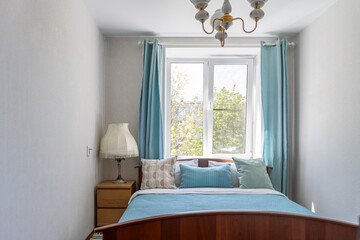 A bed in the bedroom with a blue bedspread and blue pillows. There are blue curtains on the bedroom windows. There is a bedside table with a large night lamp next to the bed