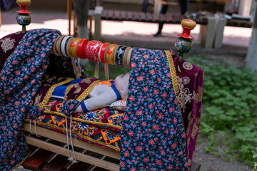 A baby doll is laying on a bed with a blanket draped over it. The bed is decorated with a colorful blanket and has a wooden frame. The scene is peaceful and calming