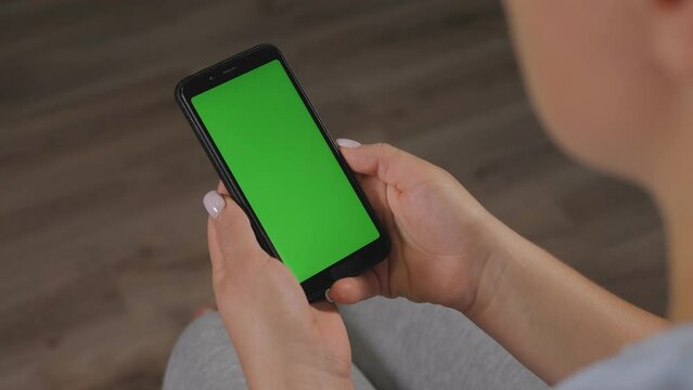 Close-up of woman's hand browsing smartphone with green screen at home. Woman at home relaxing reading on the smartphone with pre-keyed green screen.