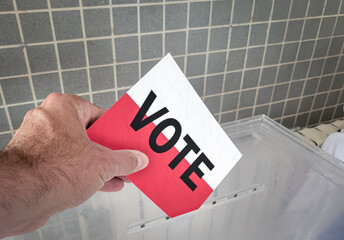 A close-up photograph captures a hand placing a voting ballot with the word VOTE printed over the...
