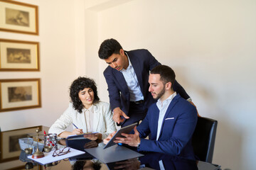 Three men in suits are sitting at a table with a tablet in front of them