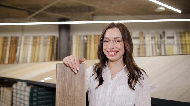 Happy young adult woman holding sample of vinyl flooring in a hardware store. Parquet board panel. Close up of a laminate flooring board. SPC flooring