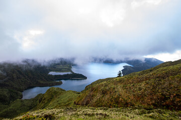 A misty view of a tranquil lake surrounded by lush green hills and low-hanging clouds.