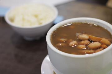 A bowl of Singapore beans soup and rice is on a table. The soup is brown and has a lot of beans in it.