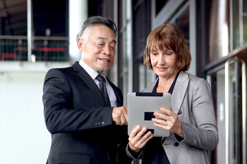 Happy Asian senior businessman in suit looking tablet discussing information with his businesswoman partner. Elderly successful boss talking about schedule with secretary at front of office workplace.