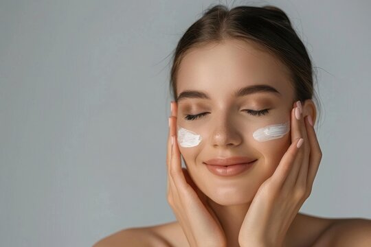 Smiling woman applying skincare cream on face against a gray background