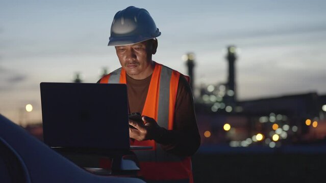 A man in an orange vest is looking at a laptop and cell phone. He is wearing a hard hat and safety glasses. The scene is set in a city at night, with a car parked nearby
