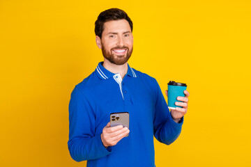 Photo portrait of handsome young guy drink coffee hold device dressed stylish blue garment isolated on yellow color background