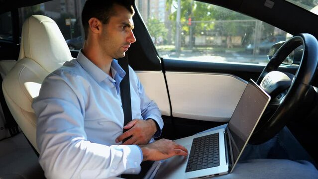 Handsome entrepreneur working on notebook while riding an autonomous self driving electric car at urban road. Male businessman typing text on laptop during riding on electrical vehicle with autopilot