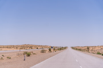 A road in the desert with a truck driving down it. The sky is clear and the sun is shining brightly