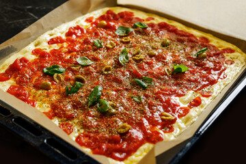 A vibrant, uncooked pizza with tomato sauce, mozzarella cheese, basil leaves, and garlic on a baking tray.