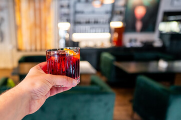 Hand holding a red cocktail with ice and an orange slice against a blurred bar background.