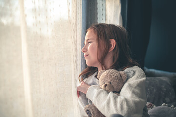 a pensive cute girl sits and looks out the window, hugging a teddy bear.