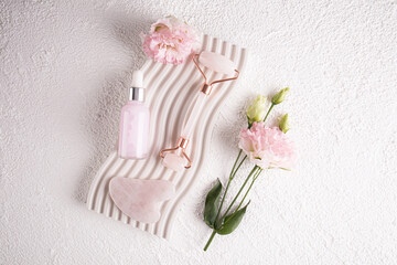 A white matte bottle of cosmetics, a rose quartz massage roller and a gua sha scraper lie on an embossed podium. Top view.