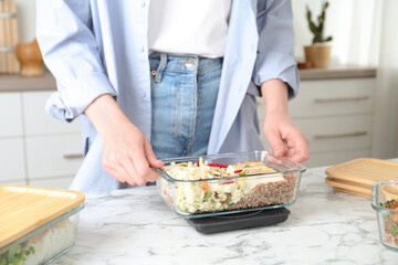 Healthy meal. Woman weighing glass container with food on kitchen scale at white marble table, closeup
