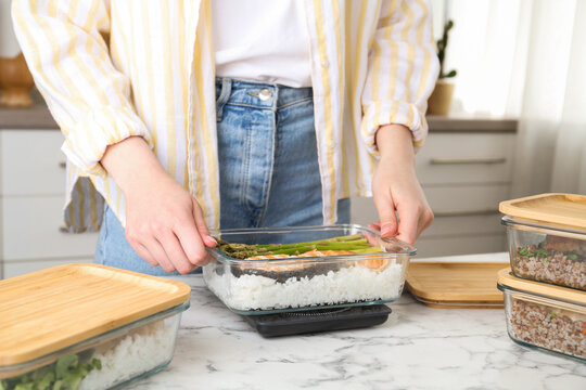 Healthy meal. Woman weighing glass container with food on kitchen scale at white marble table, closeup
