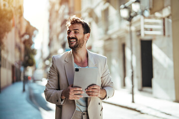 A cheerful Caucasian man in a light suit engages with a digital tablet, standing on an urban street lined with buildings basking in sunlight.