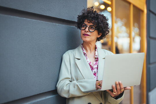 A confident Hispanic businesswoman works attentively on her laptop while standing on a city sidewalk, dressed in smart business attire.