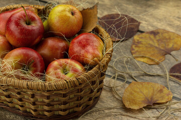 Autumn still life of apples in a wicker basket and dry leaves. Red apples in straw on a wooden background surrounded by autumn leaves. Autumn composition.