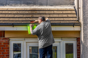 Man on ladder cleaning house roof gutters above front door removing leaves moss