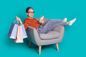 Full body photo of attractive senior woman sit armchair device shopper dressed stylish brown clothes isolated on cyan color background