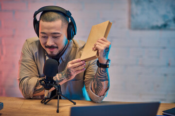 An Asian man sits in his studio, wearing headphones, and records a podcast while holding a book.