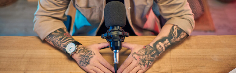 A handsome Asian man with tattooed arms sits in his podcast studio, preparing to record.