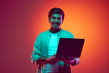 Young, attractive Indian man looks at camera while using laptop in neon light against gradient studio background. Concept of fashion and style, human emotions, study, work online.
