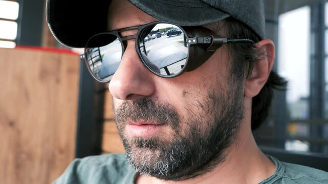 A manly man with a beard, wearing a baseball cap and steampunk style mirrored sunglasses with leather side shields, looks through the distance during the daytime in the city, slow motion closeup shot.