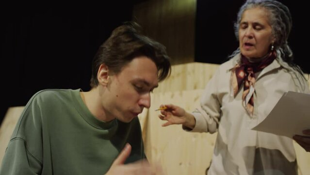 Young male actor reading script on paper and performing speech while emotional female director standing beside on stage and giving advice during theatre rehearsal