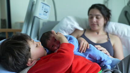 Older sibling lying on hospital bed with newborn baby, while mother watches lovingly in the background. tender family moment of bonding, warmth, and early sibling connection in a hospital setting