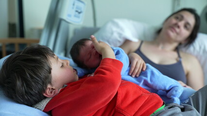 Older sibling lying on hospital bed with newborn baby, while mother watches lovingly in the background. tender family moment of bonding, warmth, and early sibling connection in a hospital setting